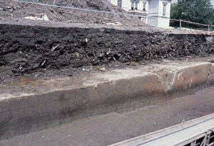 調査区の土層断面。壁面の上半分を占める、どす黒い層が、水害に伴うゴミの集積。なお一番下の薄く黒い層は弥生前期の水田。